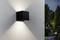 Un luminaire mural noir moderne sur un mur extérieur blanc texturé, projetant des motifs de lumière symétriques. Le jardin et les arbres sont visibles en arrière-plan.