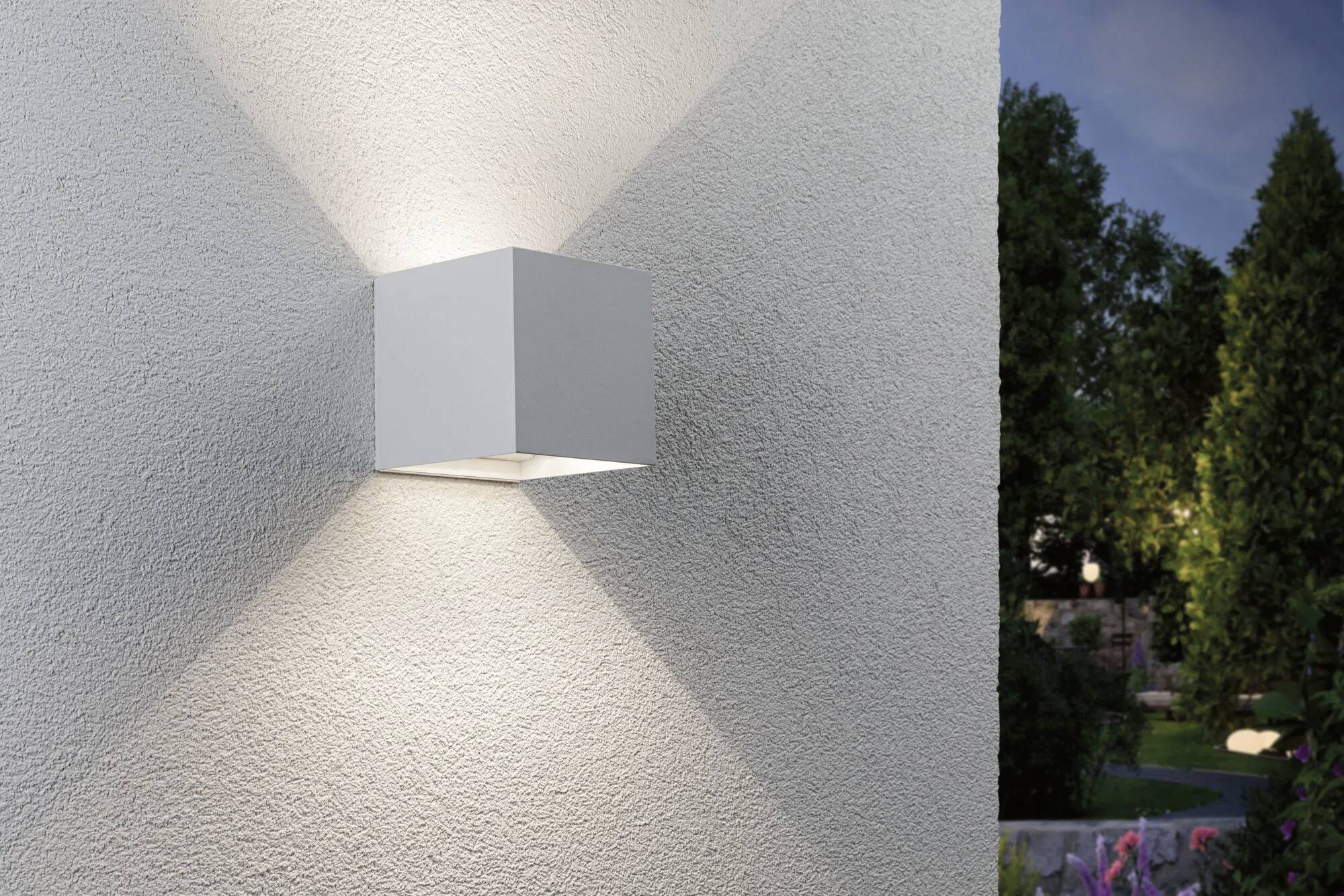 Applique murale cubique lumineuse fixée sur un mur extérieur texturé, projetant des ombres symétriques formant une forme en V. Des arbres sont visibles en arrière-plan.
