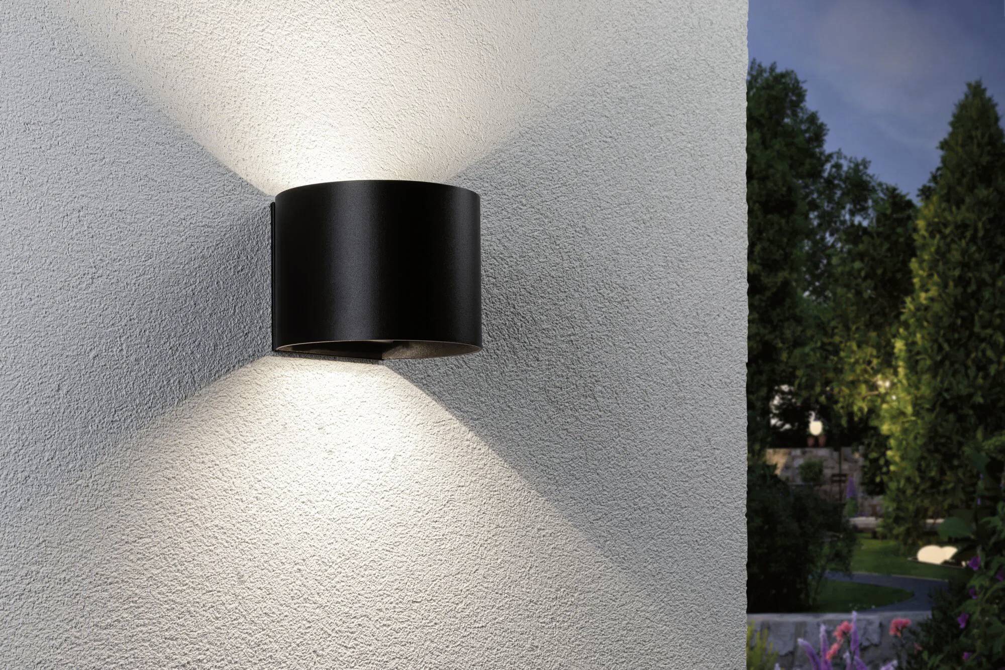 Un luminaire mural noir fixé à l'extérieur sur un mur blanc texturé, projetant de la lumière vers le haut et vers le bas, avec un jardin visible en arrière-plan.