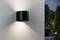Un luminaire mural noir fixé à l'extérieur sur un mur blanc texturé, projetant de la lumière vers le haut et vers le bas, avec un jardin visible en arrière-plan.