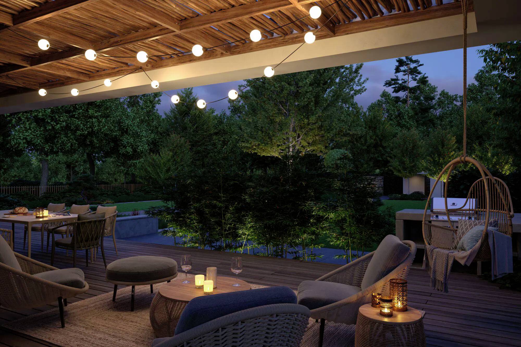 Terrasse extérieure la nuit avec des guirlandes lumineuses, des sièges et une table dressée pour un dîner. Des arbres en arrière-plan créent une atmosphère chaleureuse et intime.