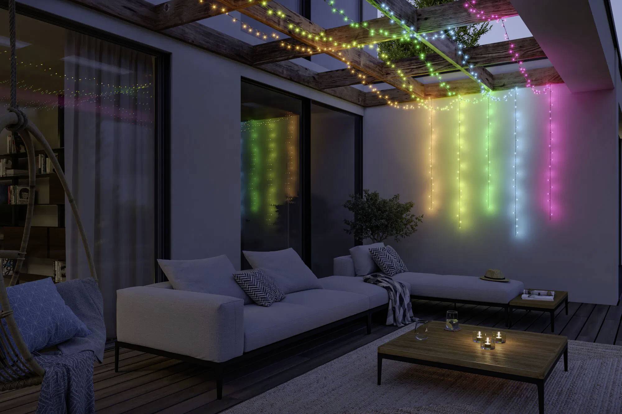 Terrasse extérieure de nuit avec deux canapés, une table basse et une pergola ornée de guirlandes lumineuses multicolores qui diffusent un éclairage d'ambiance.
