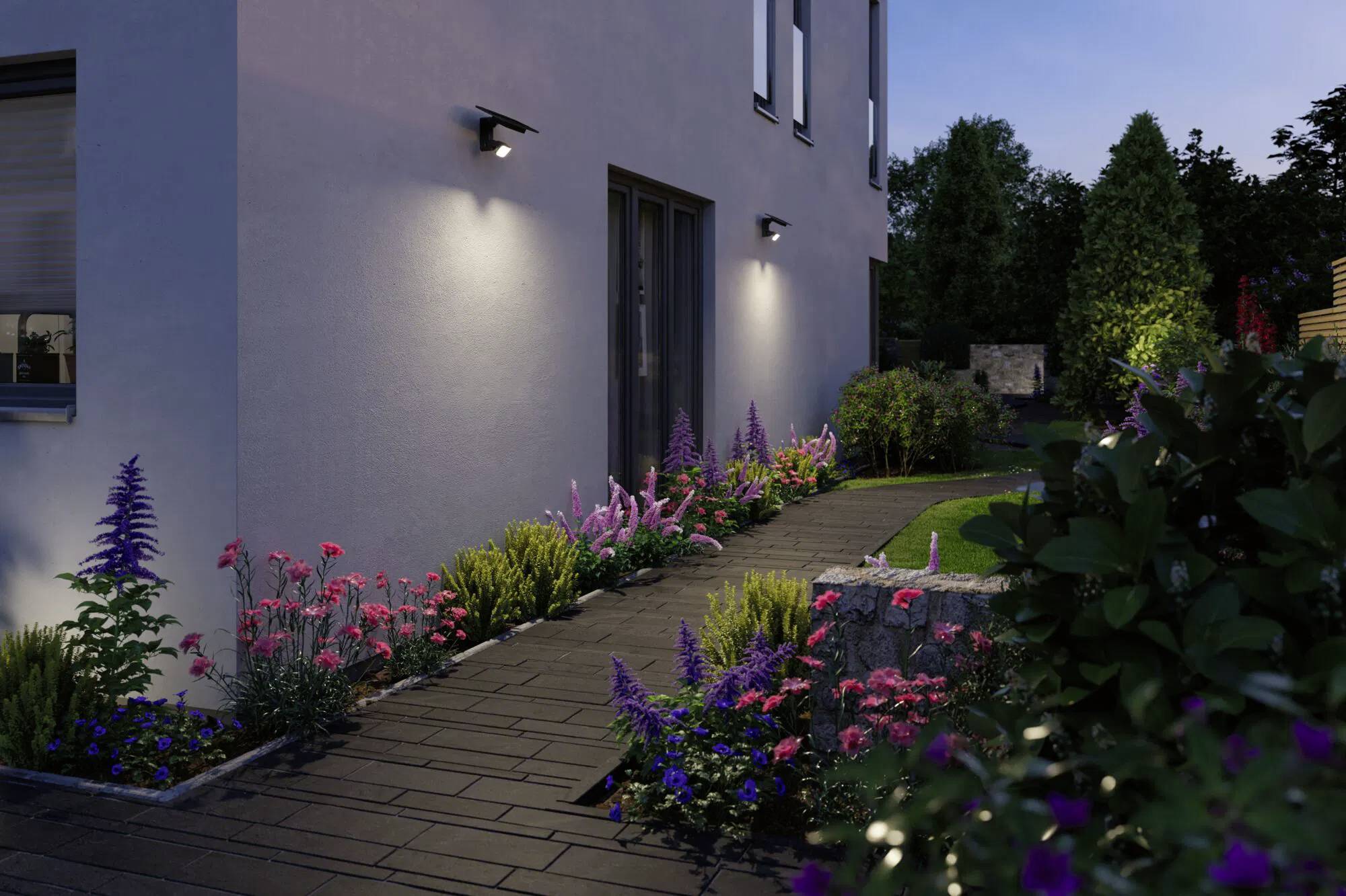 Sentier bordé de fleurs colorées et d'arbustes menant à l'entrée d'une maison, éclairé par des luminaires muraux extérieurs pendant la soirée.