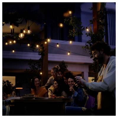 Un groupe de personnes profitant d'un rassemblement autour d'une table extérieure, chaleureusement éclairé par des guirlandes lumineuses, avec un homme debout à proximité tenant une bouteille.