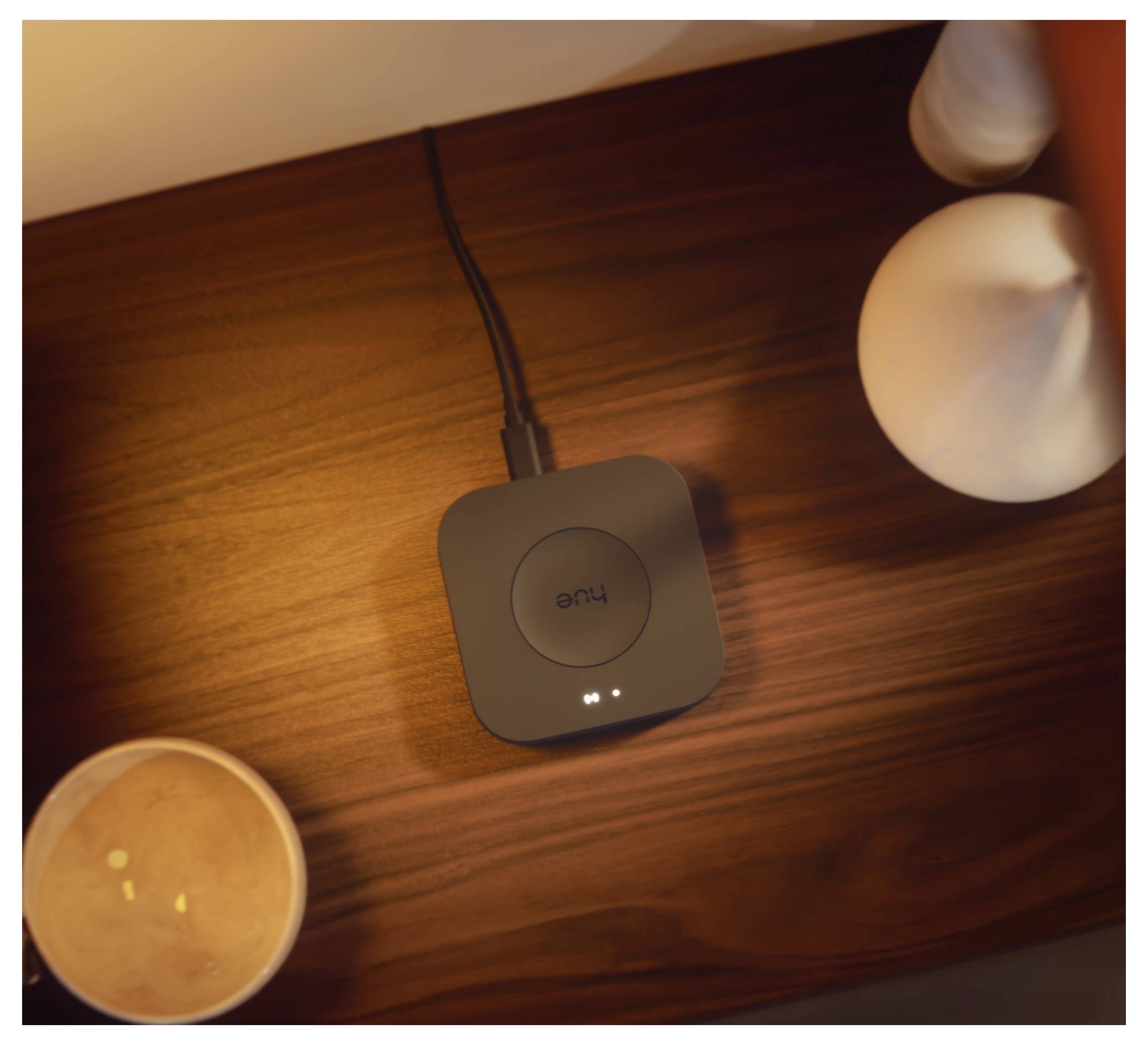 Un chargeur sans fil posé sur une surface en bois avec un voyant lumineux, à côté d'objets décoratifs tels qu'une bougie et un petit vase blanc.