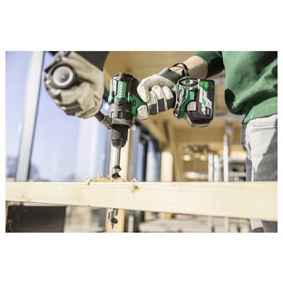 Une personne utilise une perceuse sans fil verte pour percer un trou dans une planche de bois dans un atelier lumineux, portant des gants de sécurité.