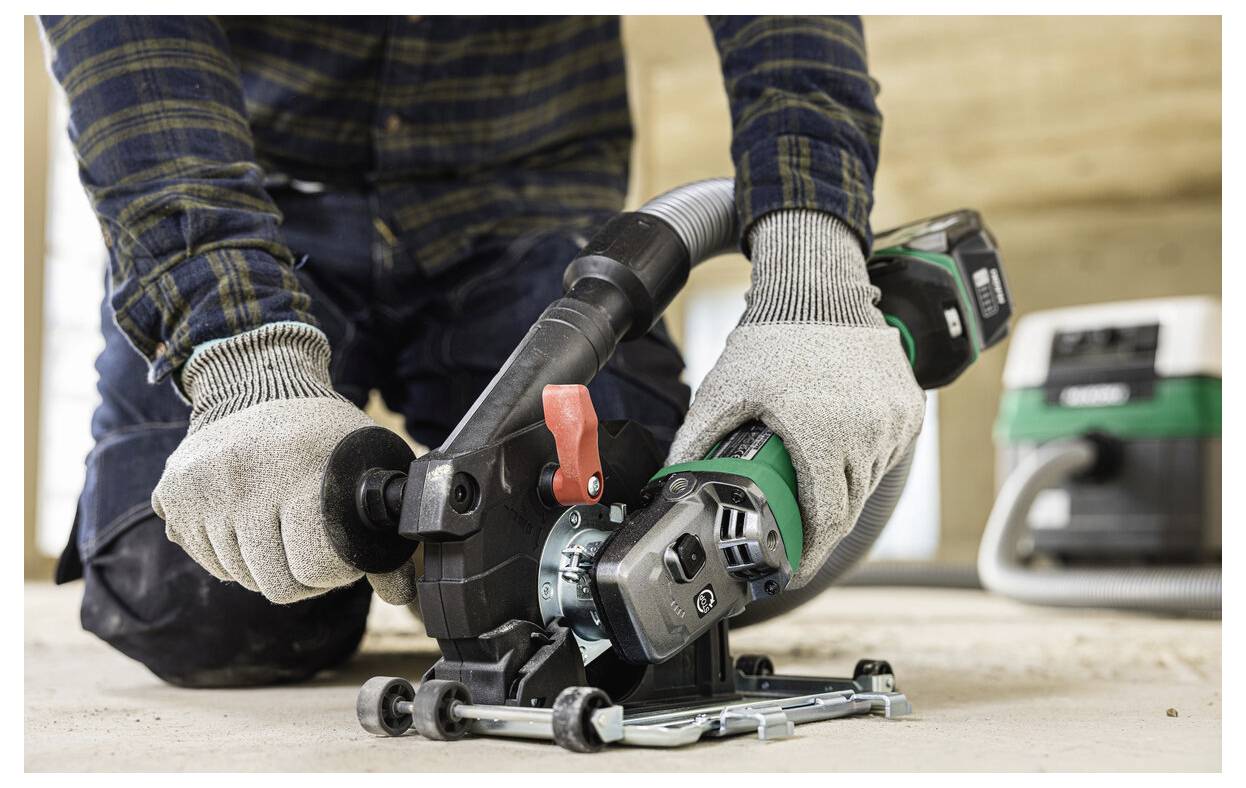 Une personne portant des gants utilise un outil électrique équipé d'un système d'aspiration des poussières. L'outil est utilisé sur une surface en bois dans un environnement de construction.