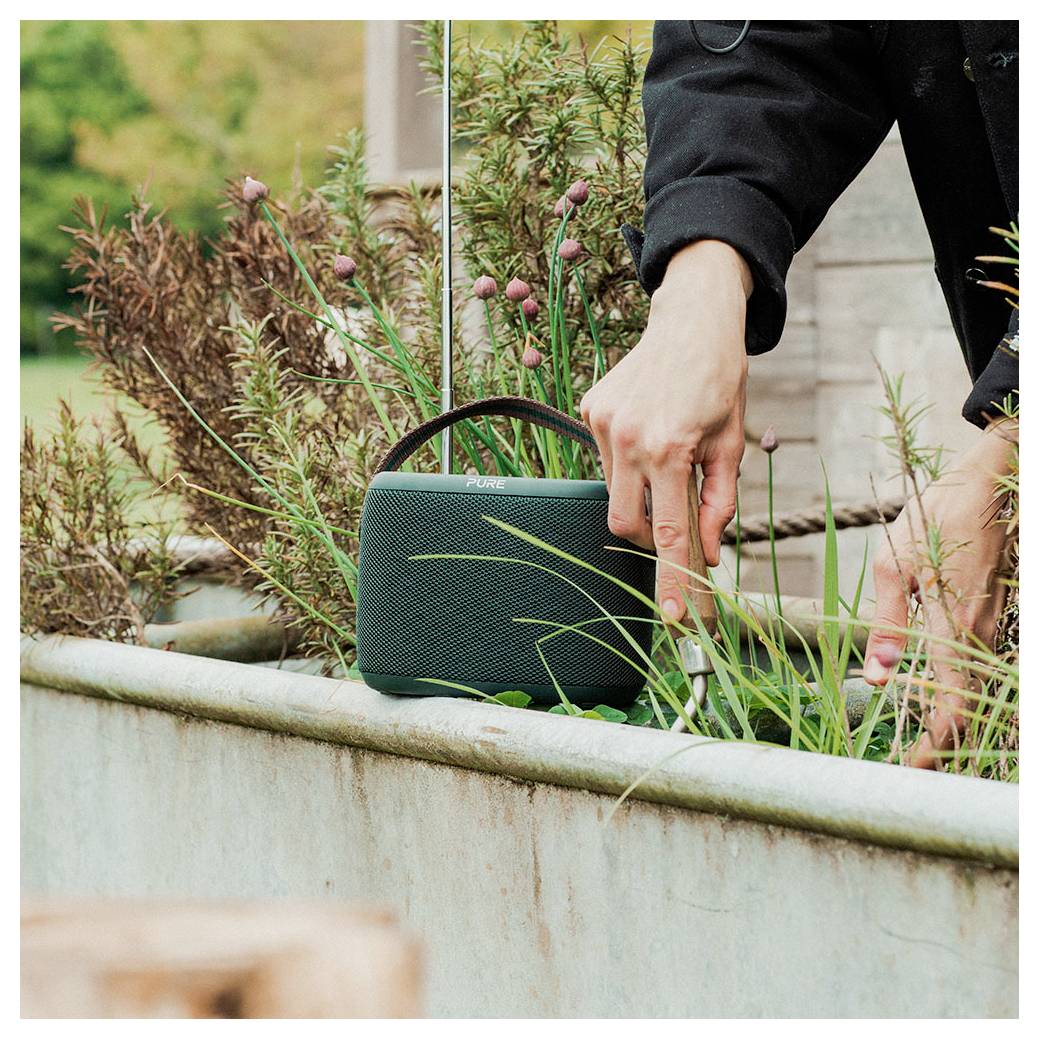 Une personne soigne des plantes dans un jardin tout en tenant une enceinte portable verte, suggérant des loisirs en plein air ou un travail accompagné de musique.