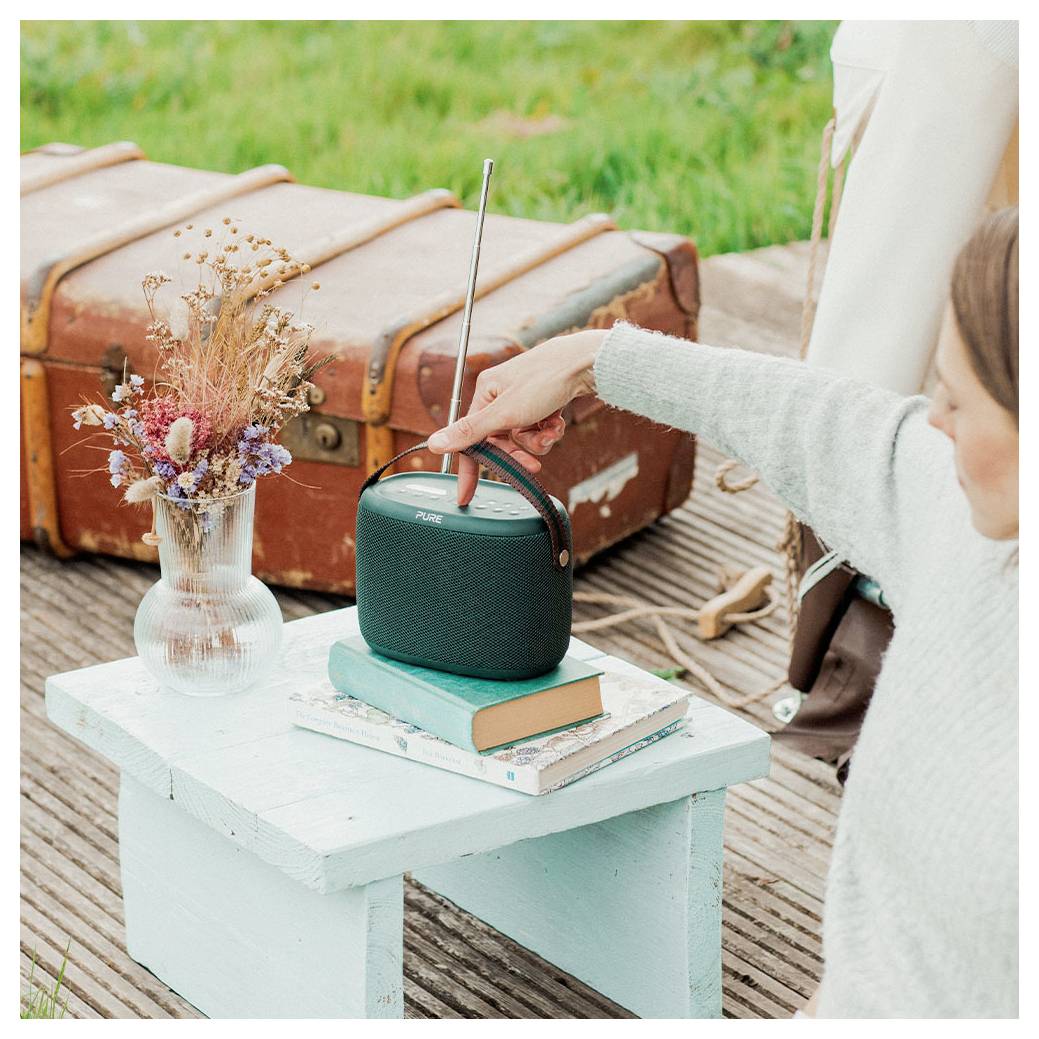 Une personne règle une radio portable équipée d'une antenne, placée sur une petite table supportant des livres et un vase de fleurs séchées, installée à l'extérieur près de valises.