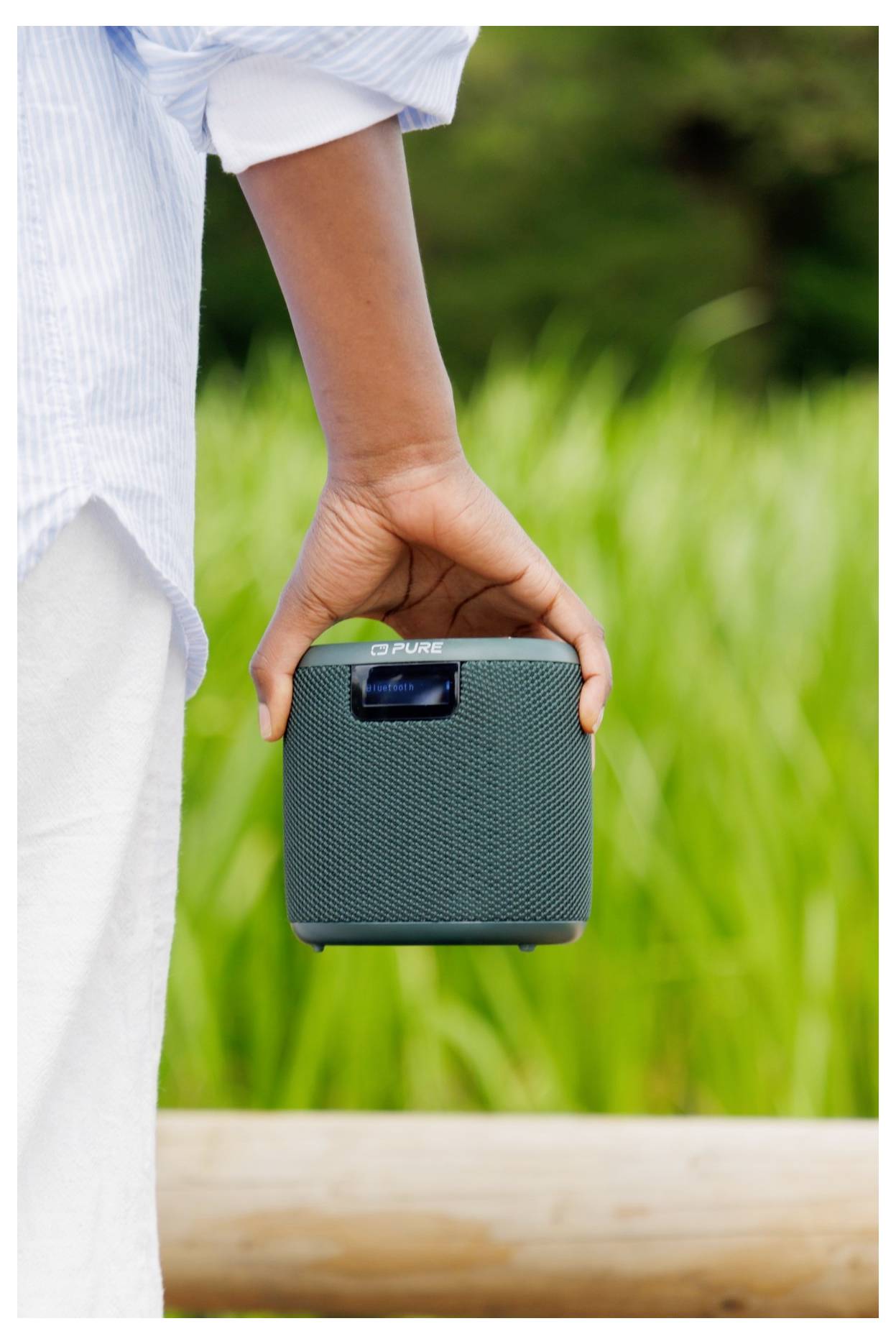 Une personne tenant un haut-parleur portable à l'extérieur, avec de l'herbe verte en arrière-plan.