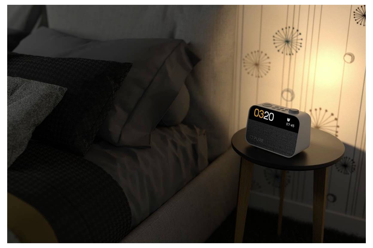 Un réveil numérique affichant '03:20' sur une table de chevet dans une chambre faiblement éclairée, à côté d'un lit avec des oreillers et une lampe chaleureuse.