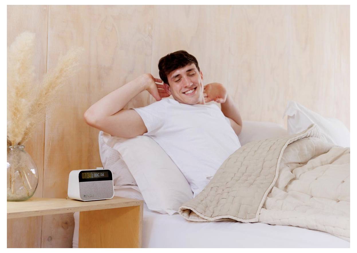 Une personne allongée dans un lit s'étire avec un sourire, à côté d'une table de chevet sur laquelle un réveil indique '7:00 du matin'. La chambre est doucement éclairée.