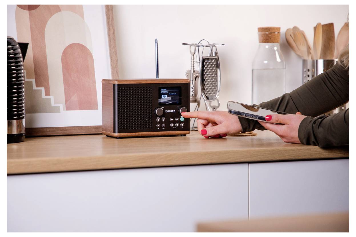 Une personne ajuste une radio numérique posée sur un comptoir de cuisine en bois tout en tenant un smartphone ; des ustensiles de cuisine sont visibles en arrière-plan.