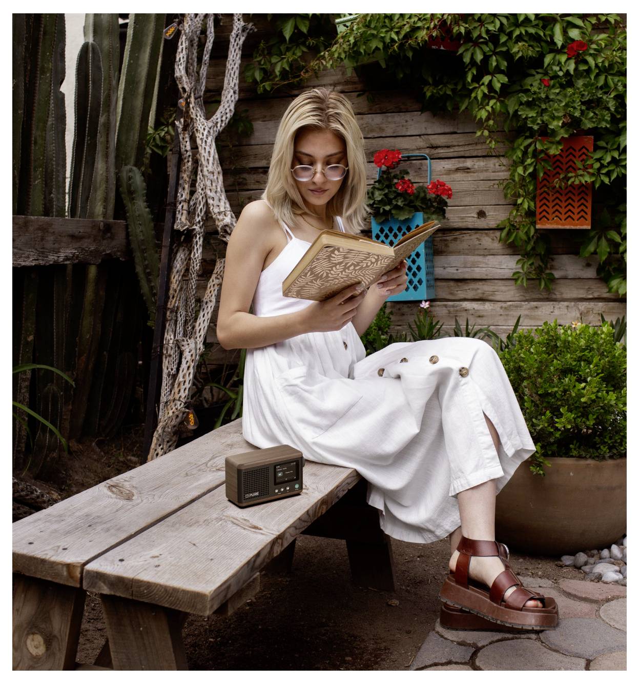Une femme portant une robe blanche et des lunettes de soleil lit un livre assise sur un banc en bois dans un jardin avec des cactus et des lanternes colorées.