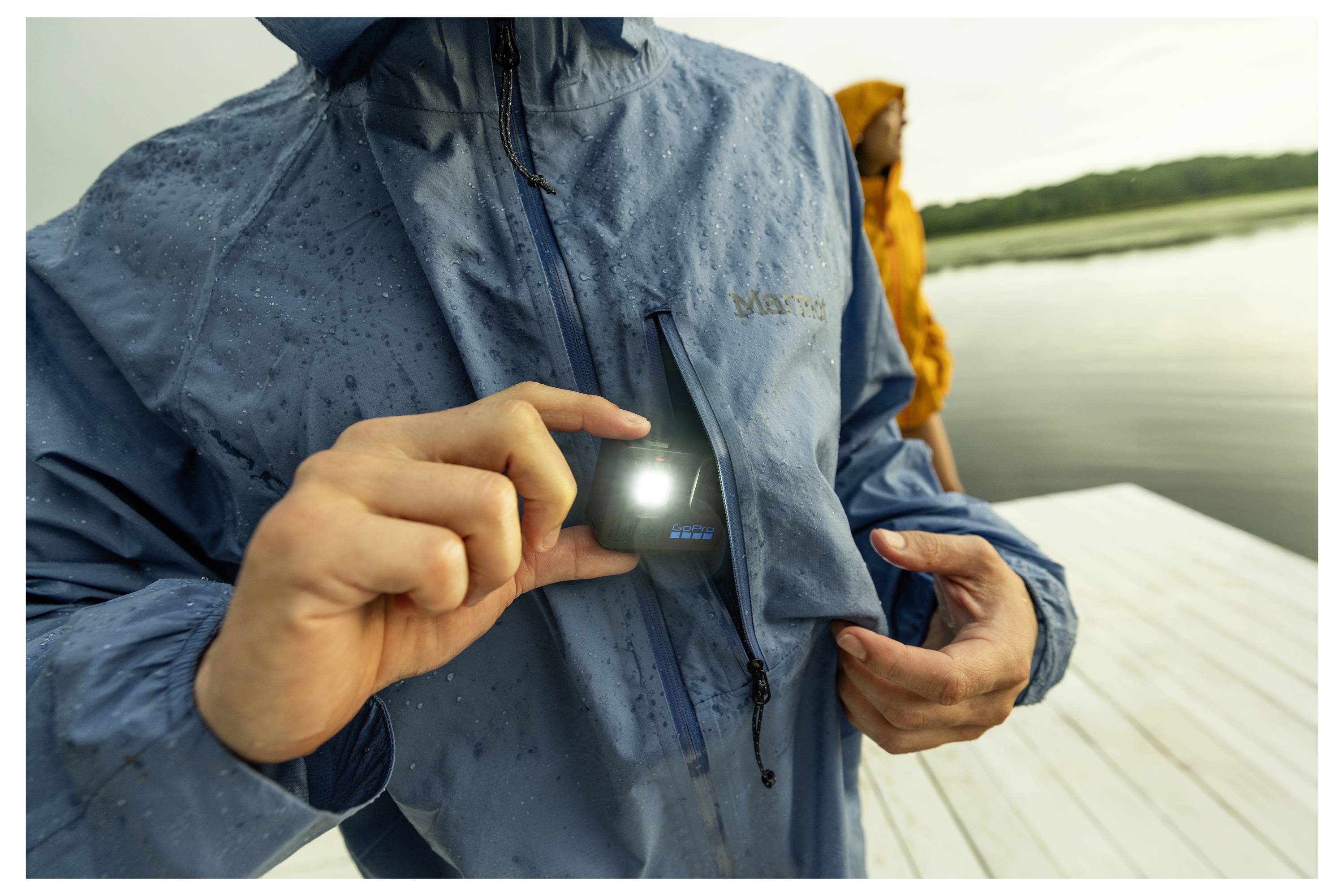 Une personne portant un imperméable tient un petit appareil LED illuminé près de la poche de poitrine. Une autre personne portant une veste jaune se tient près de l'eau.