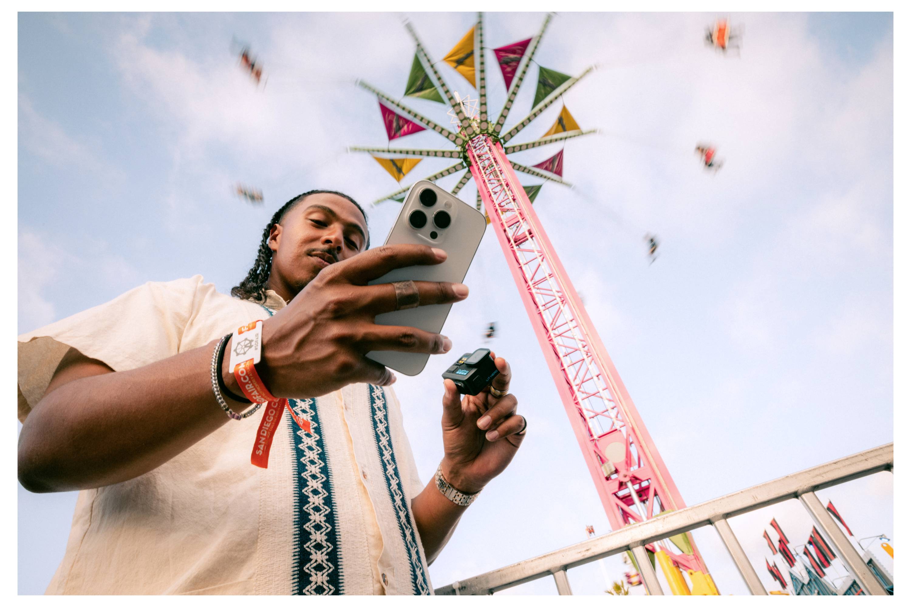 Une personne utilisant un smartphone se tient devant une grande attraction de parc d'attractions colorée, avec des gens balançant en mouvement sur un ciel bleu.