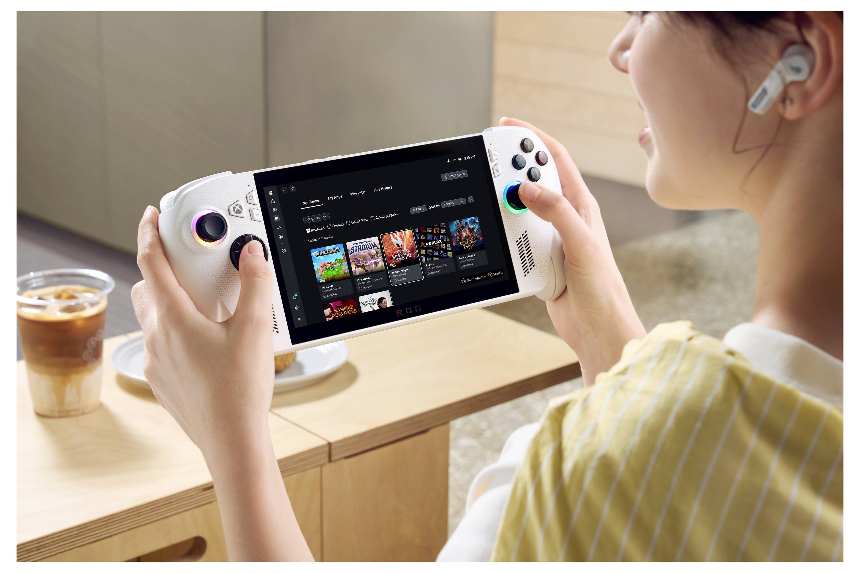 Une personne utilise un appareil de jeu portable avec un écran affichant une bibliothèque de jeux vidéo. Une boisson est posée sur la table à côté d'elle.