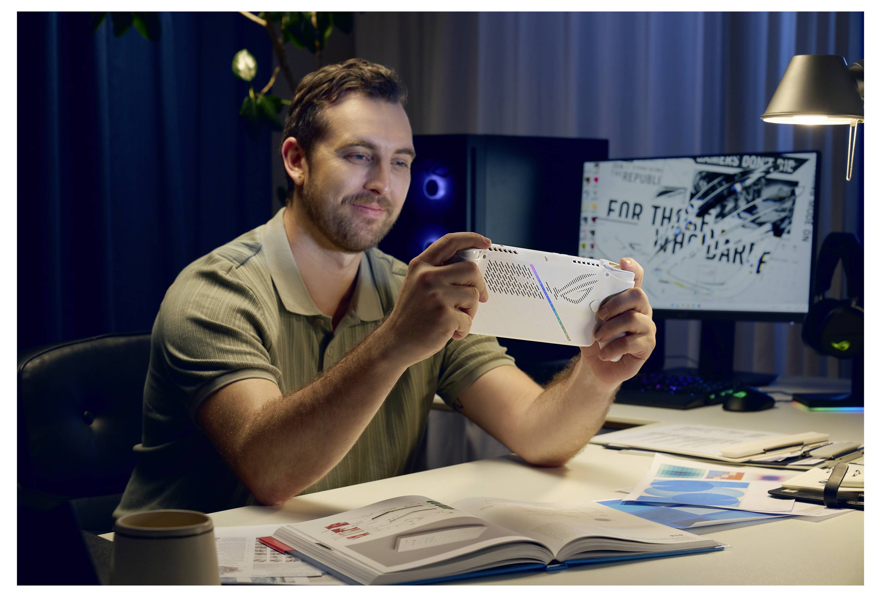Une personne est assise à un bureau, souriant à un appareil. Derrière elle, un écran d'ordinateur allumé affiche un contenu indistinct dans un environnement de bureau.