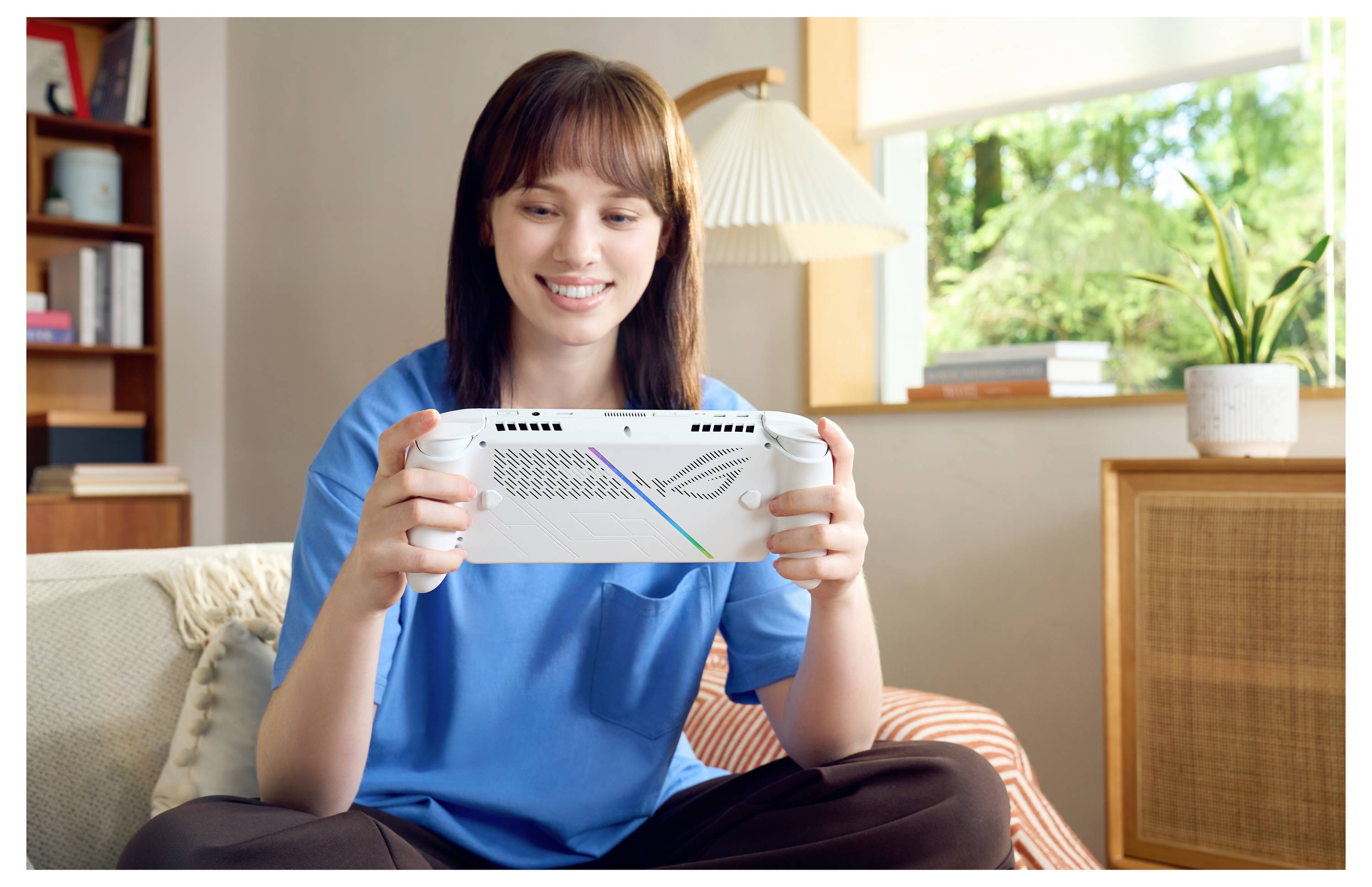 Une personne assise sur un canapé, souriant tout en utilisant un appareil de jeu portable, avec une lampe et une fenêtre montrant de la verdure en arrière-plan.