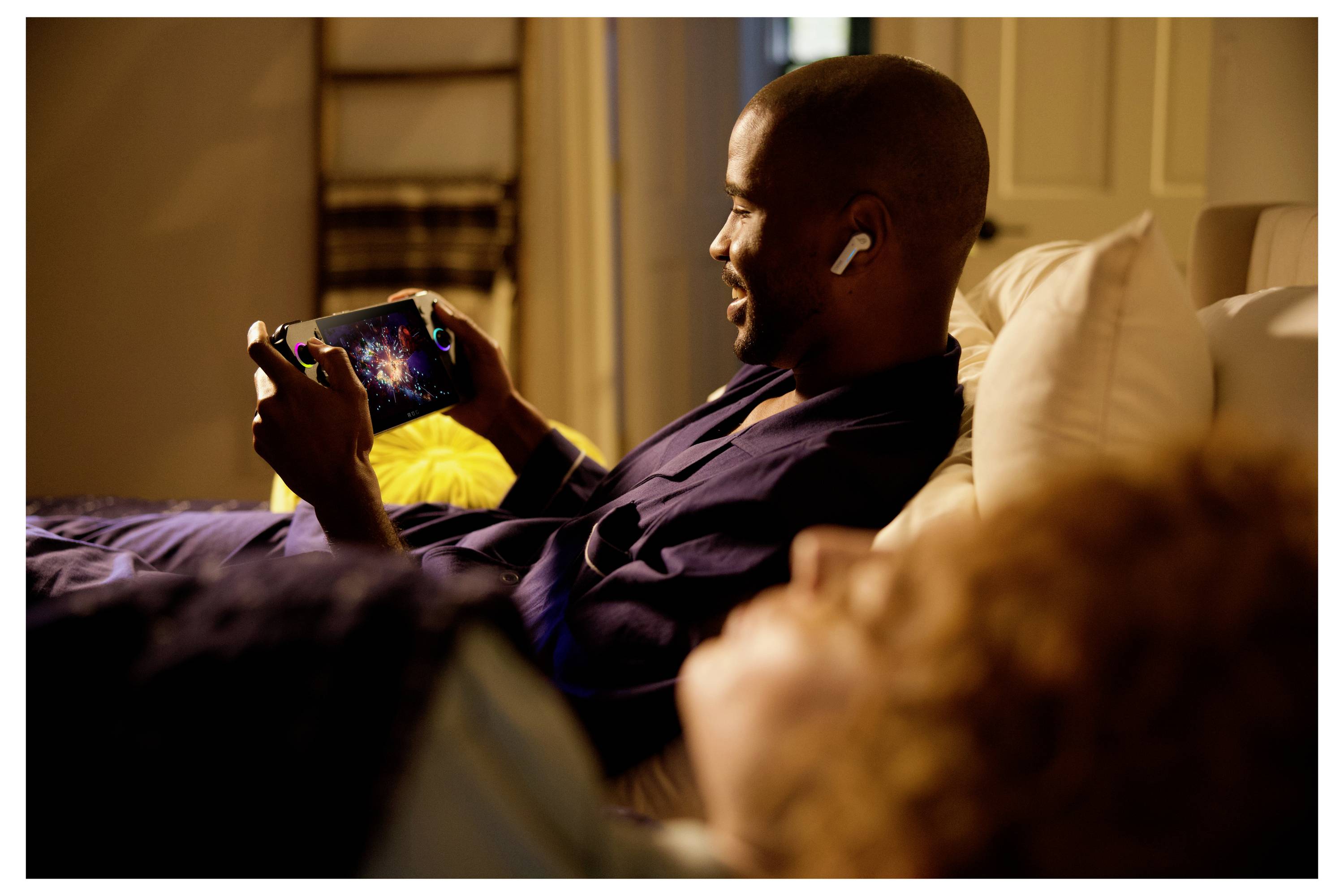 Une personne portant des écouteurs est en train de jouer à un jeu sur un appareil portable tout en étant allongée sur un lit, une autre personne se reposant à côté d'elle.