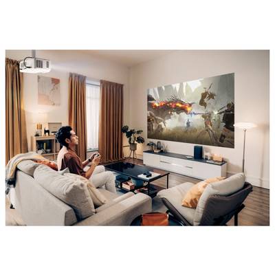 Une personne est assise sur un canapé, jouant à un jeu vidéo sur un grand écran de projection qui affiche une scène de bataille fantastique dans un salon moderne.