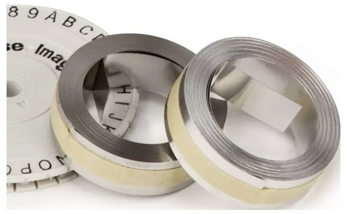 Deux pièces métalliques circulaires, chacune contenant une bande enroulée à l'intérieur, sont posées à côté d'un disque rond portant des marquages alphanumériques. Ces éléments sont probablement des composants d'un dispositif mécanique.