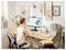 Une personne assise à un bureau, travaillant sur un ordinateur. L'écran affiche un document avec des formes abstraites. L'espace est décoré de plantes et d'œuvres encadrées.