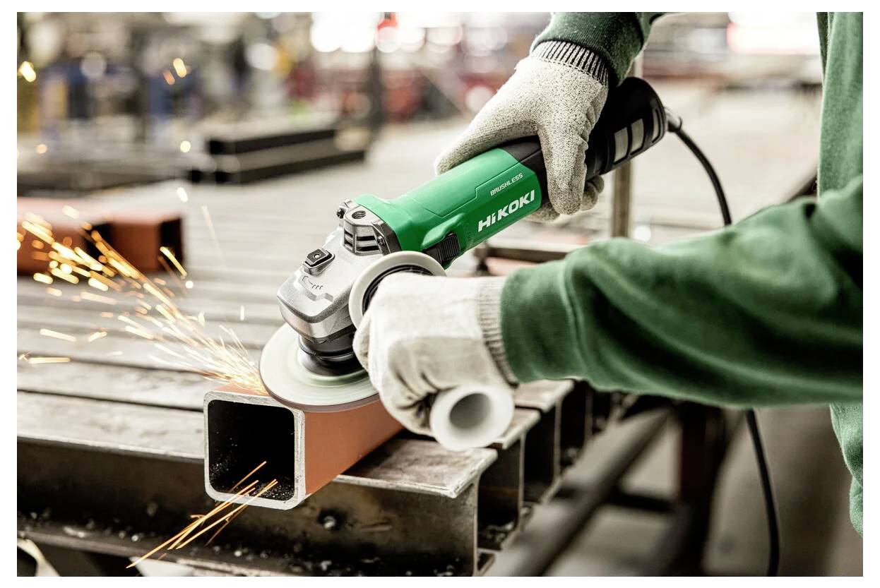 Une personne portant des gants utilise une meuleuse d'angle verte Hitachi pour lisser les bords d'une poutre métallique, avec des étincelles jaillissant du point de contact.