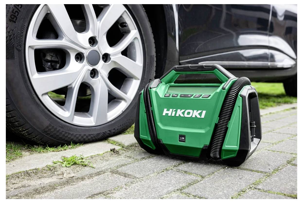 Aspirateur de marque 'HiKOKI' de couleur verte posé à côté d'un pneu de voiture sur une allée. L'aspirateur est doté d'une poignée et de tuyaux attachés.