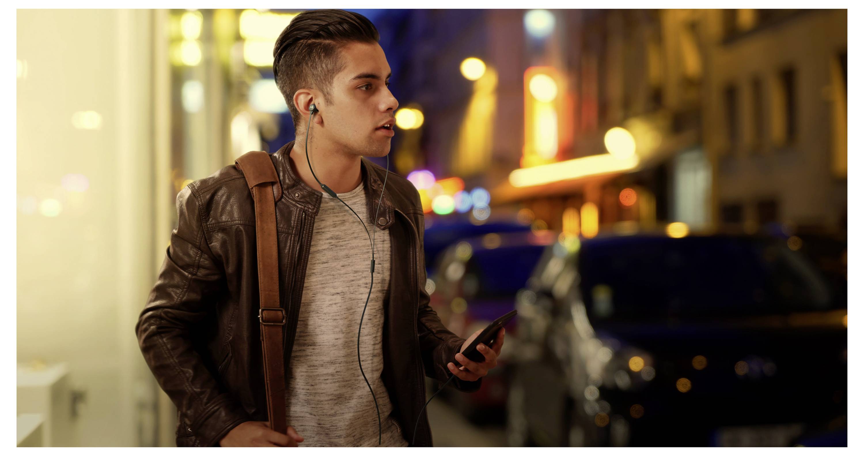 Une personne portant des écouteurs et tenant un téléphone marche le long d'une rue de ville la nuit, entourée de lumières floues et de voitures garées.