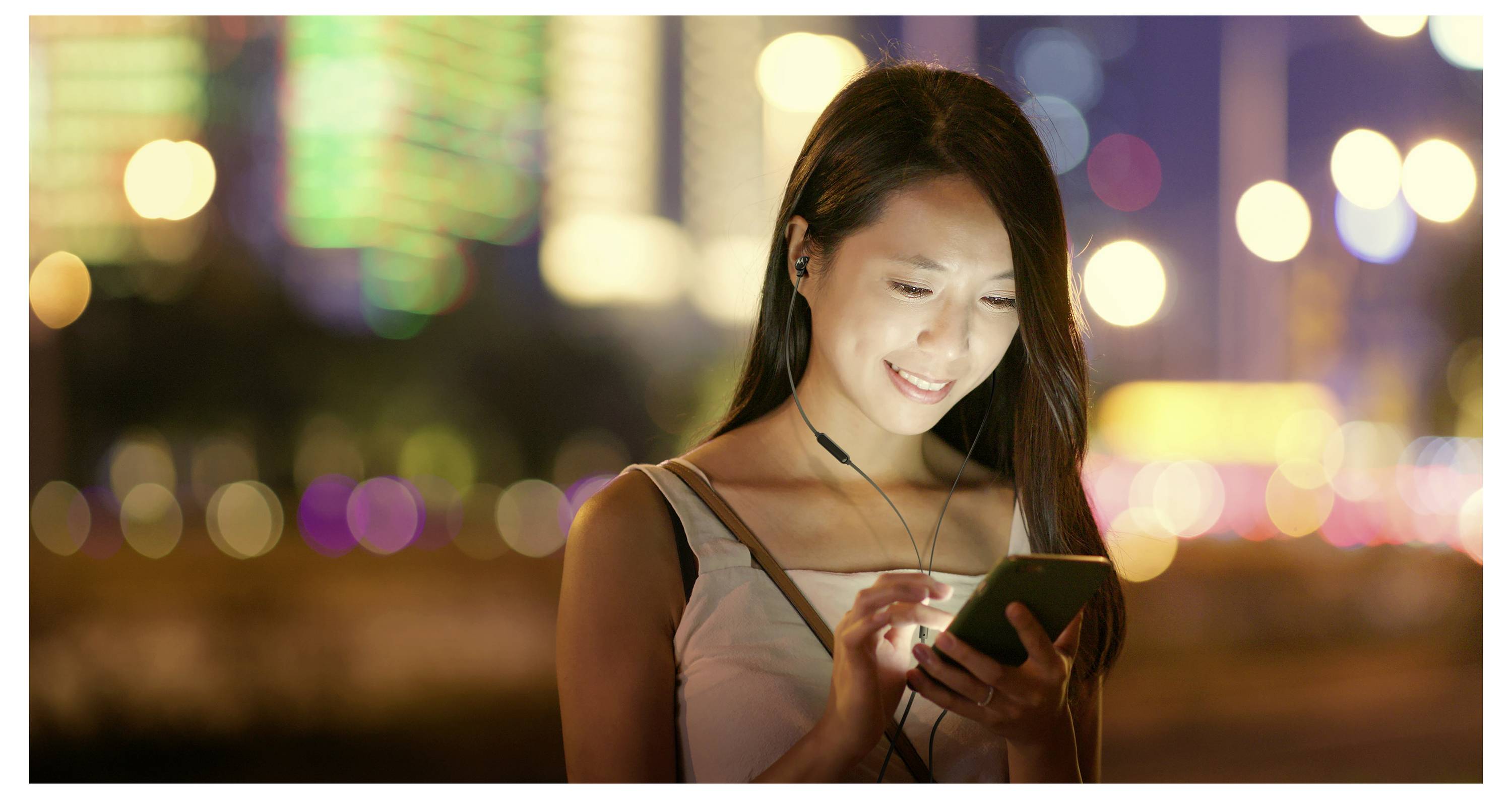 Une personne sourit en regardant un smartphone la nuit, avec des lumières de ville floues en arrière-plan.