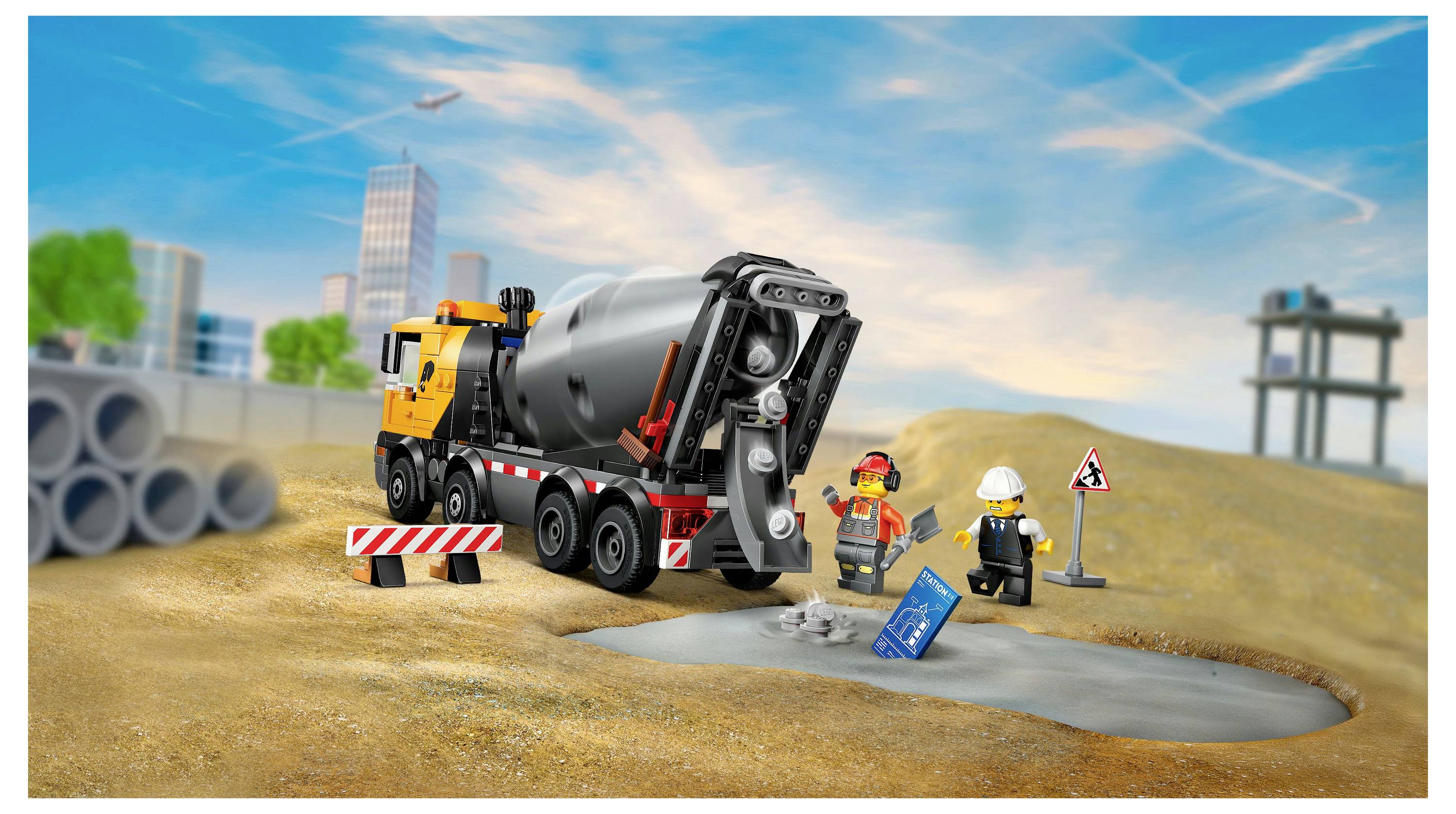 Un camion malaxeur de jouet sur un chantier de construction avec deux ouvriers miniatures. Un ouvrier tient un plan. Divers éléments de construction sont représentés.