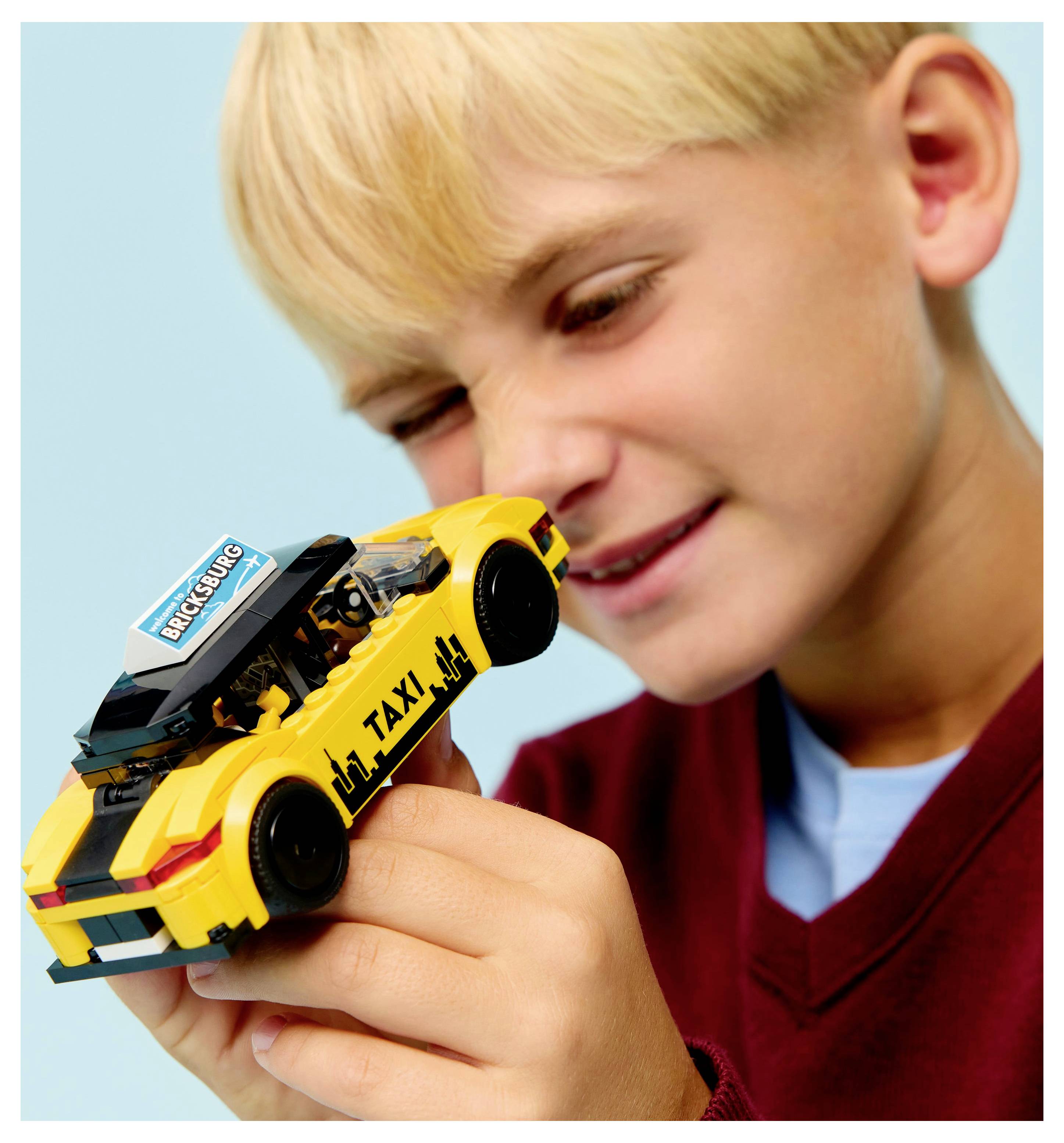 Un enfant examine un taxi jouet jaune avec l'inscription 'TAXI' sur le côté et un panneau 'Worcester' sur le toit, sur un fond bleu clair.