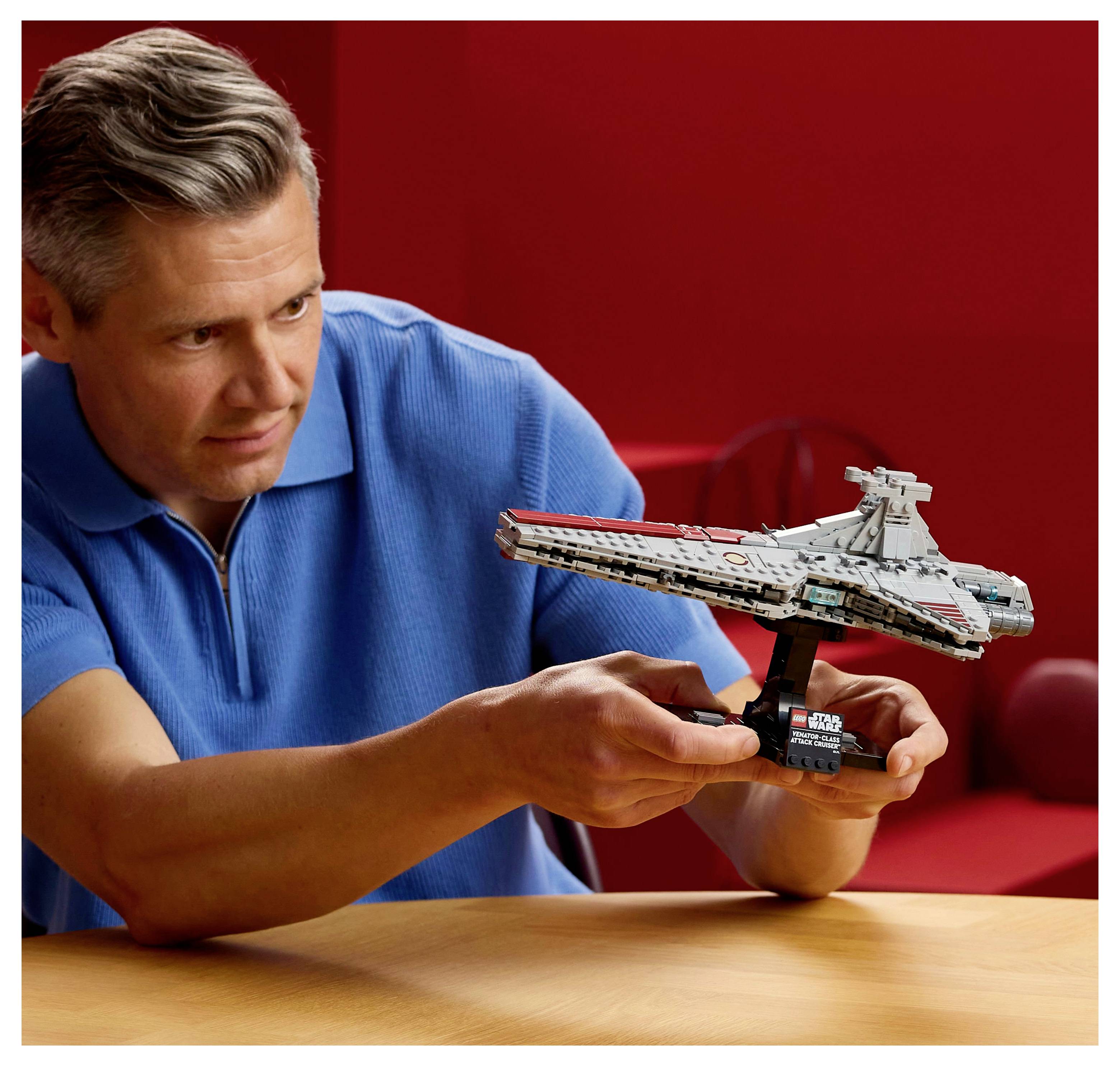 Une personne portant une chemise bleue tient un modèle détaillé d'un vaisseau spatial sur un support, sur un fond rouge. Le modèle ressemble à un vaisseau spatial de fiction.