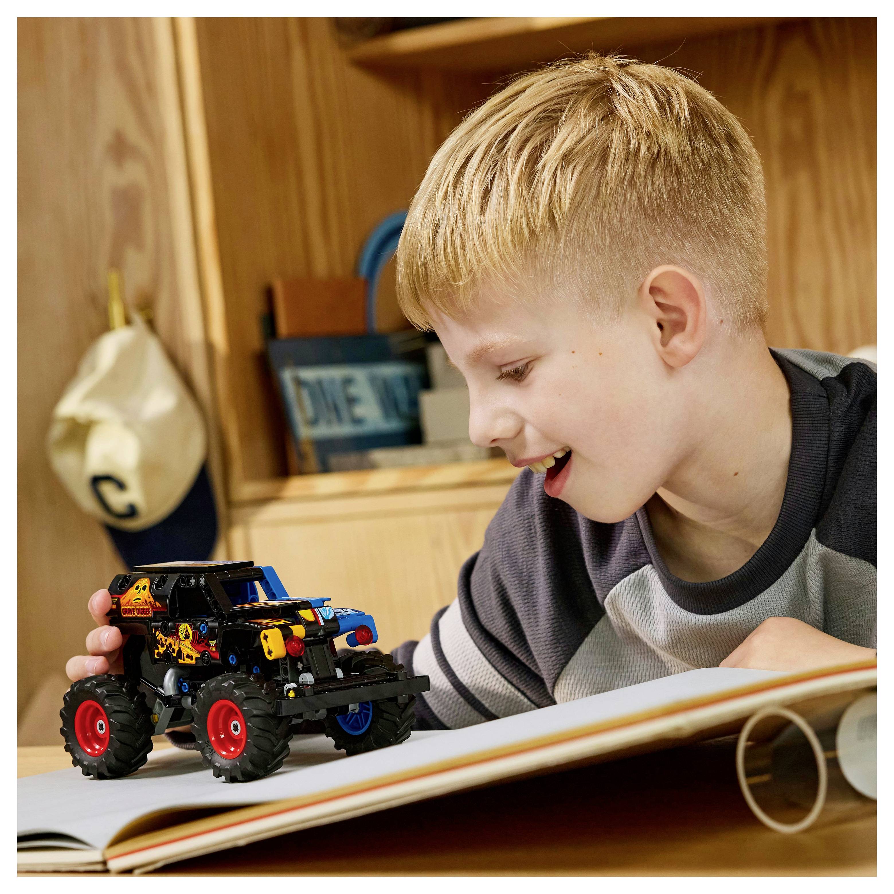 Un enfant sourit en jouant avec une voiture miniature sur un livre, dans une pièce en bois douillette. Un chapeau portant la lettre « C » est visible en arrière-plan.