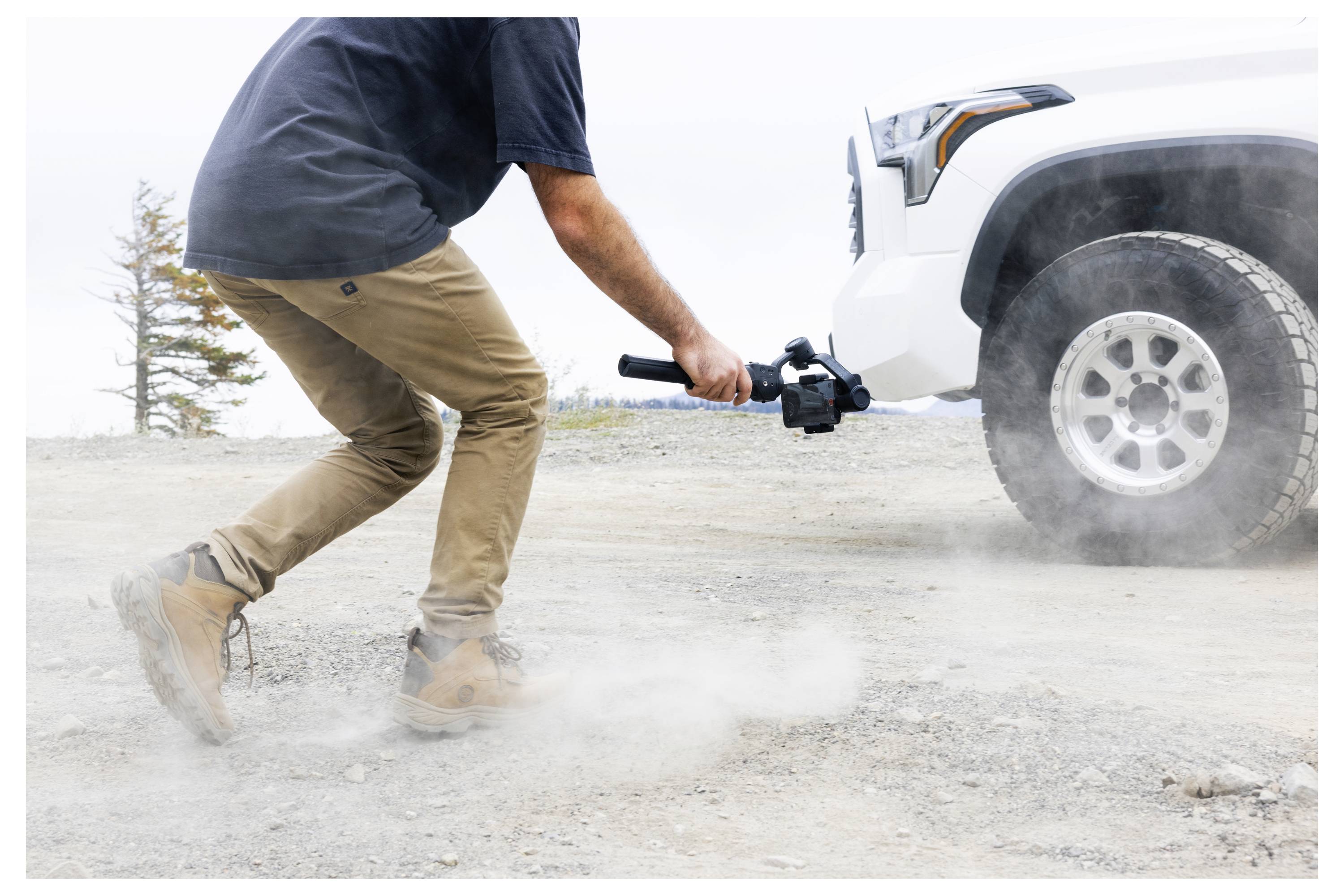 Une personne filme des images près du sol à l'aide d'un stabilisateur de caméra portable à proximité d'un véhicule garé dans un environnement extérieur poussiéreux.
