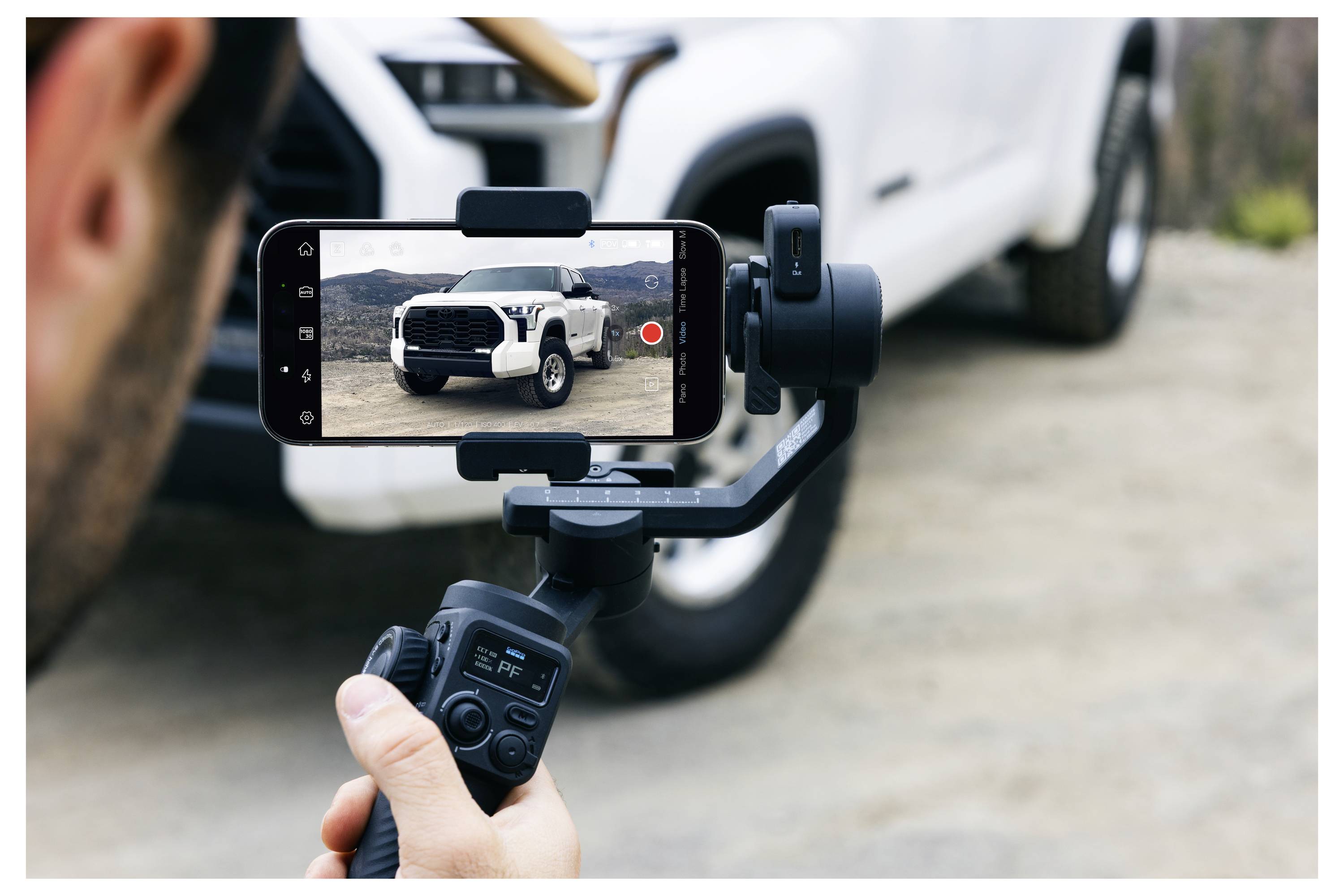 Une personne utilise un smartphone fixé sur un stabilisateur pour filmer un SUV blanc garé sur une allée.