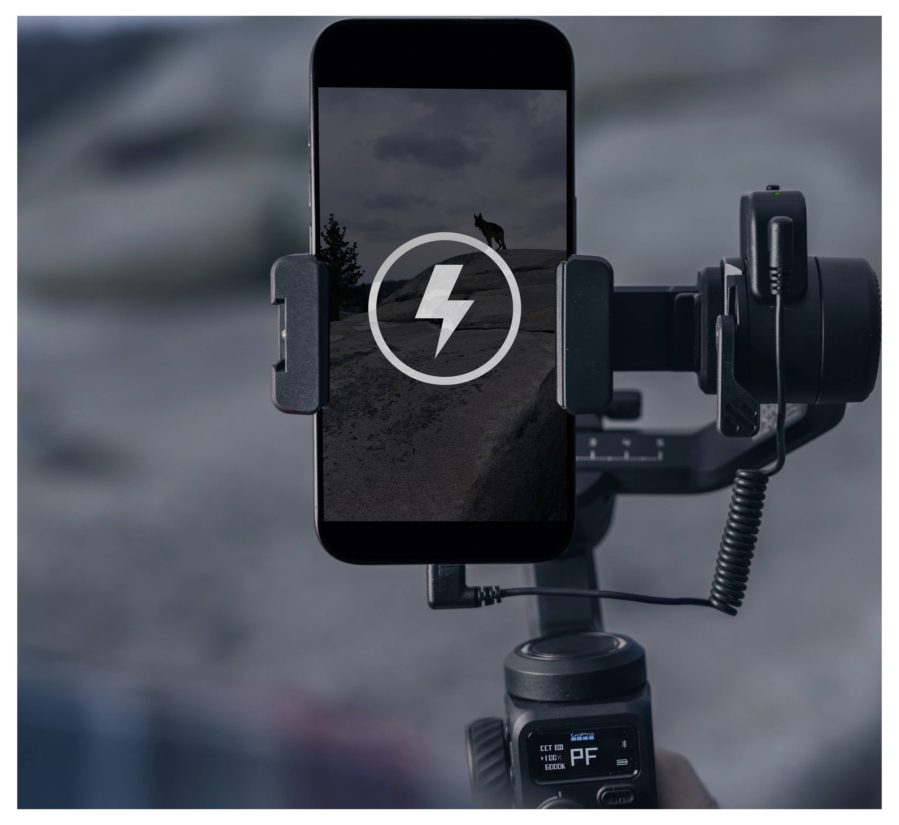 Un smartphone monté sur un cardan affiche une icône de charge sur une image de paysage avec une silhouette lointaine sur une colline.