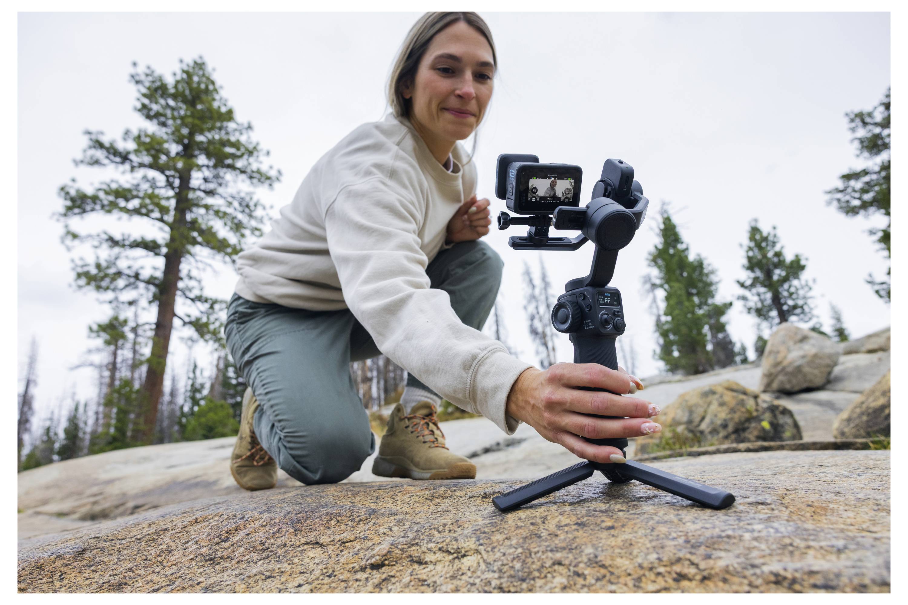 Une personne s'accroupit sur une surface rocheuse, ajustant un appareil photo portable sur un trépied, avec des arbres en arrière-plan.
