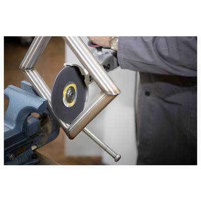 Une personne portant une tenue grise utilise un outil électrique pour couper un tuyau métallique maintenu en place par un étau, dans un environnement d'atelier.