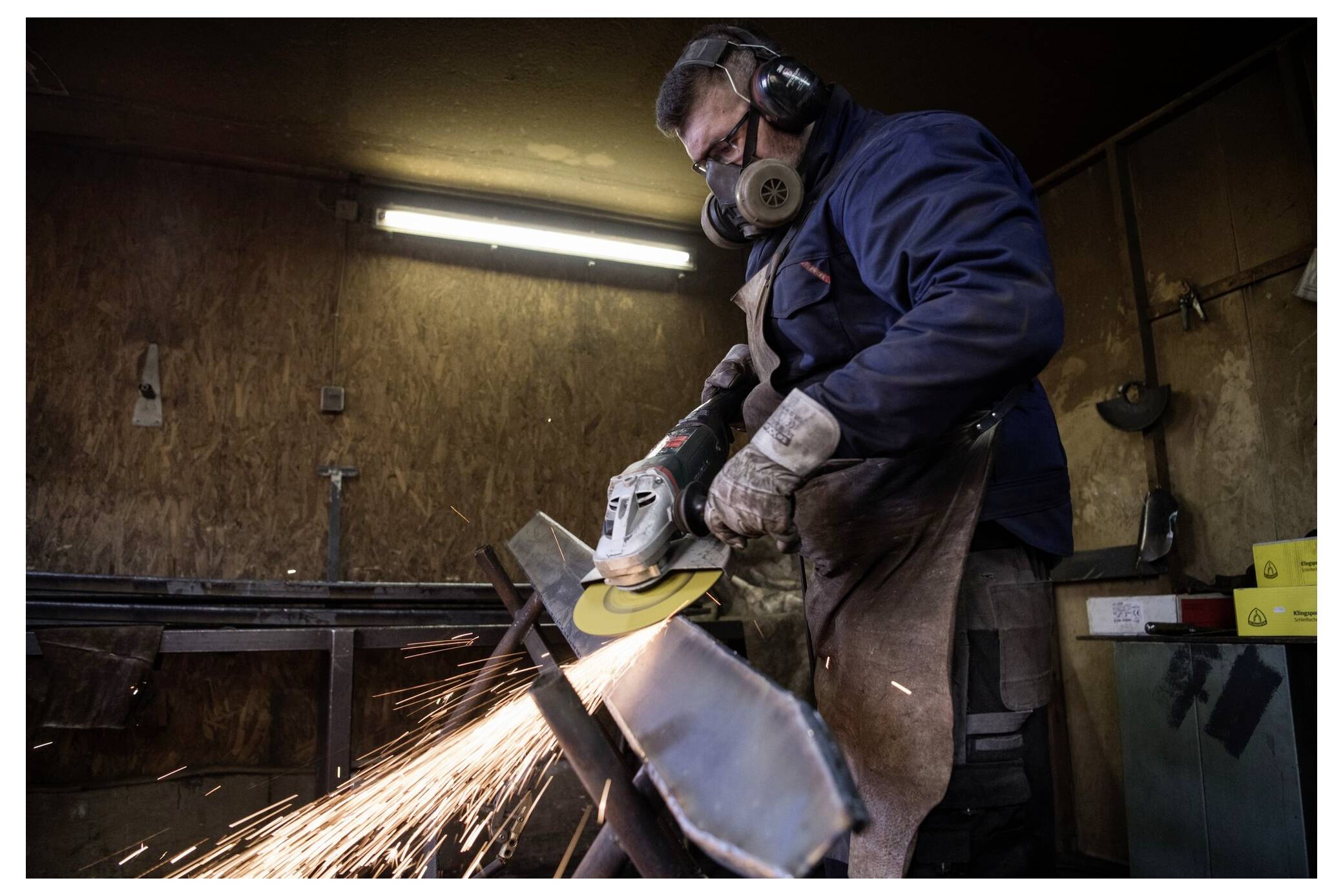 Une personne portant un équipement de protection utilise une meuleuse d'angle sur un objet métallique, créant des étincelles dans un environnement d'atelier.