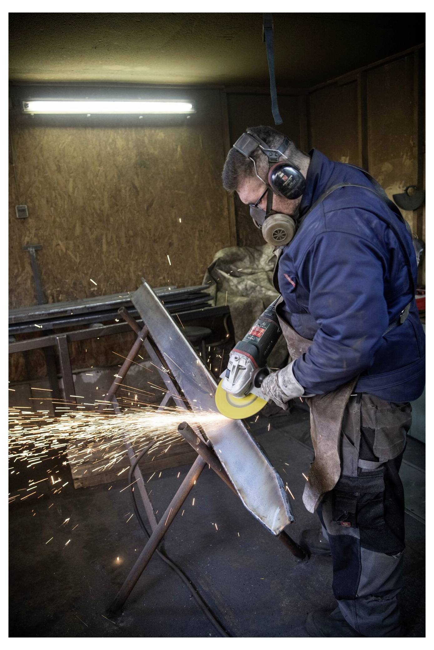 Une personne portant un équipement de protection utilise un outil de meulage sur une pièce métallique, provoquant des étincelles dans un environnement d'atelier.