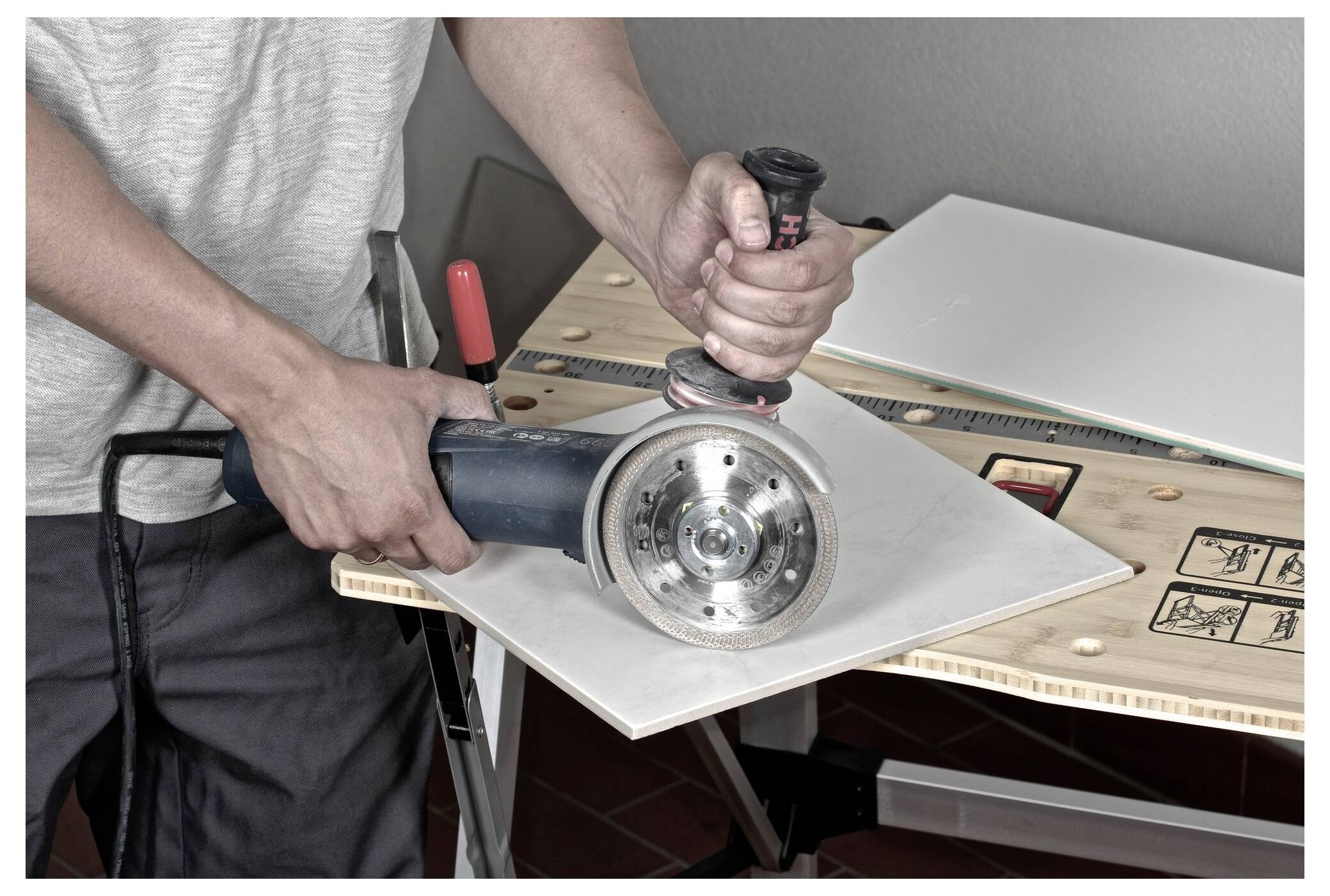 Une personne utilise une meuleuse d'angle pour couper une grande dalle en céramique blanche sur un établi. Des instructions de sécurité sont visibles sur l'établi.