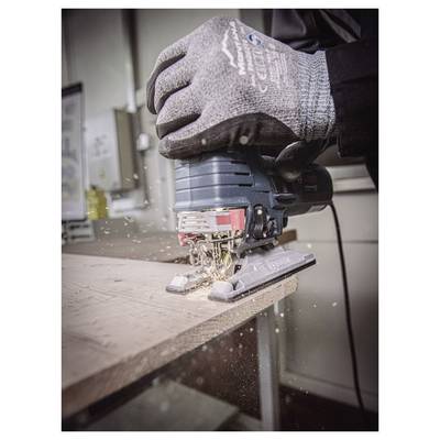 Une personne portant des gants utilise une scie sauteuse pour couper une planche en bois, avec des copeaux de sciure se dispersant. Le cadre semble être un atelier.