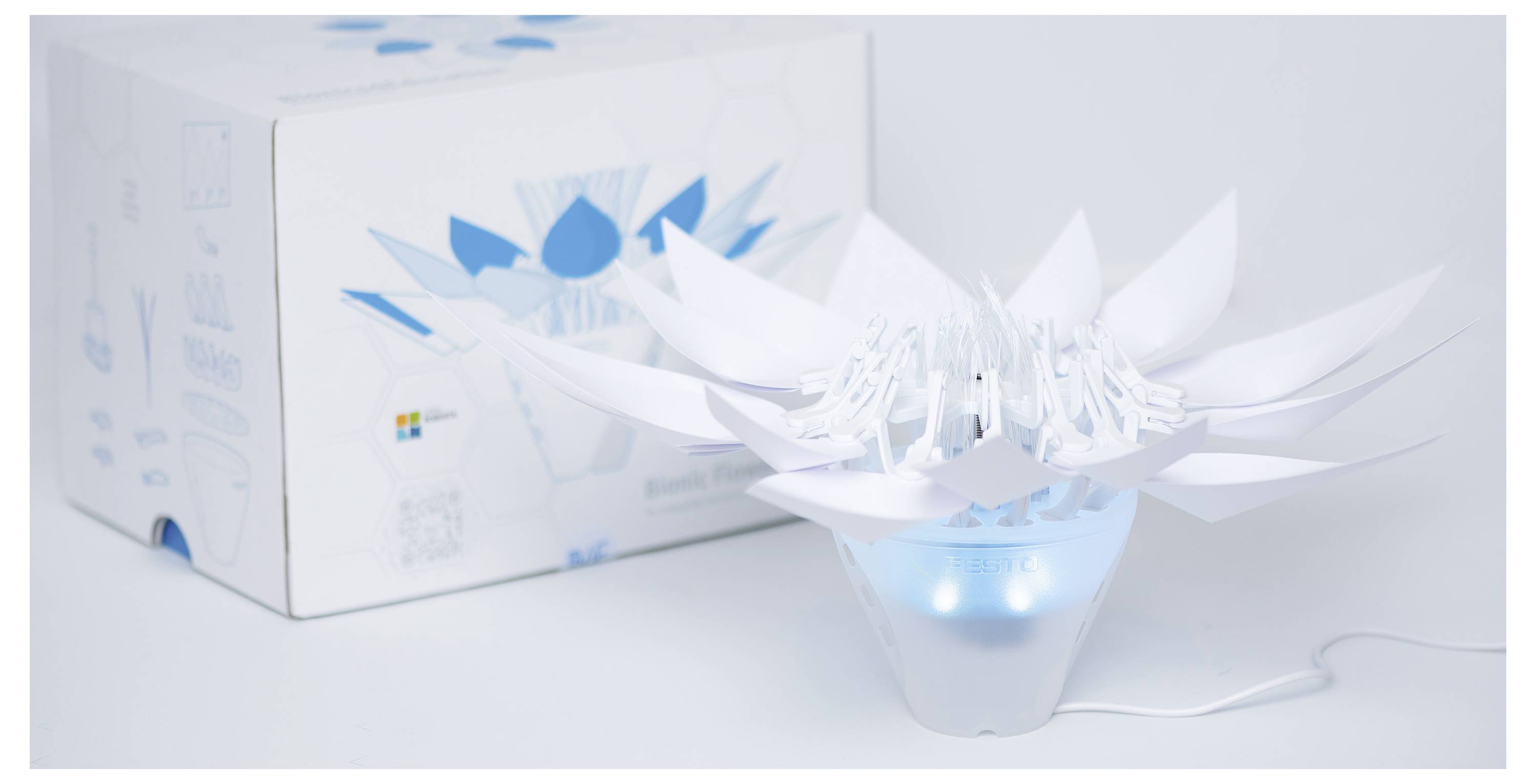Une lampe décorative blanche et bleue en forme de fleur, avec des pétales déployés, à côté de sa boîte d'emballage, sur un fond blanc.