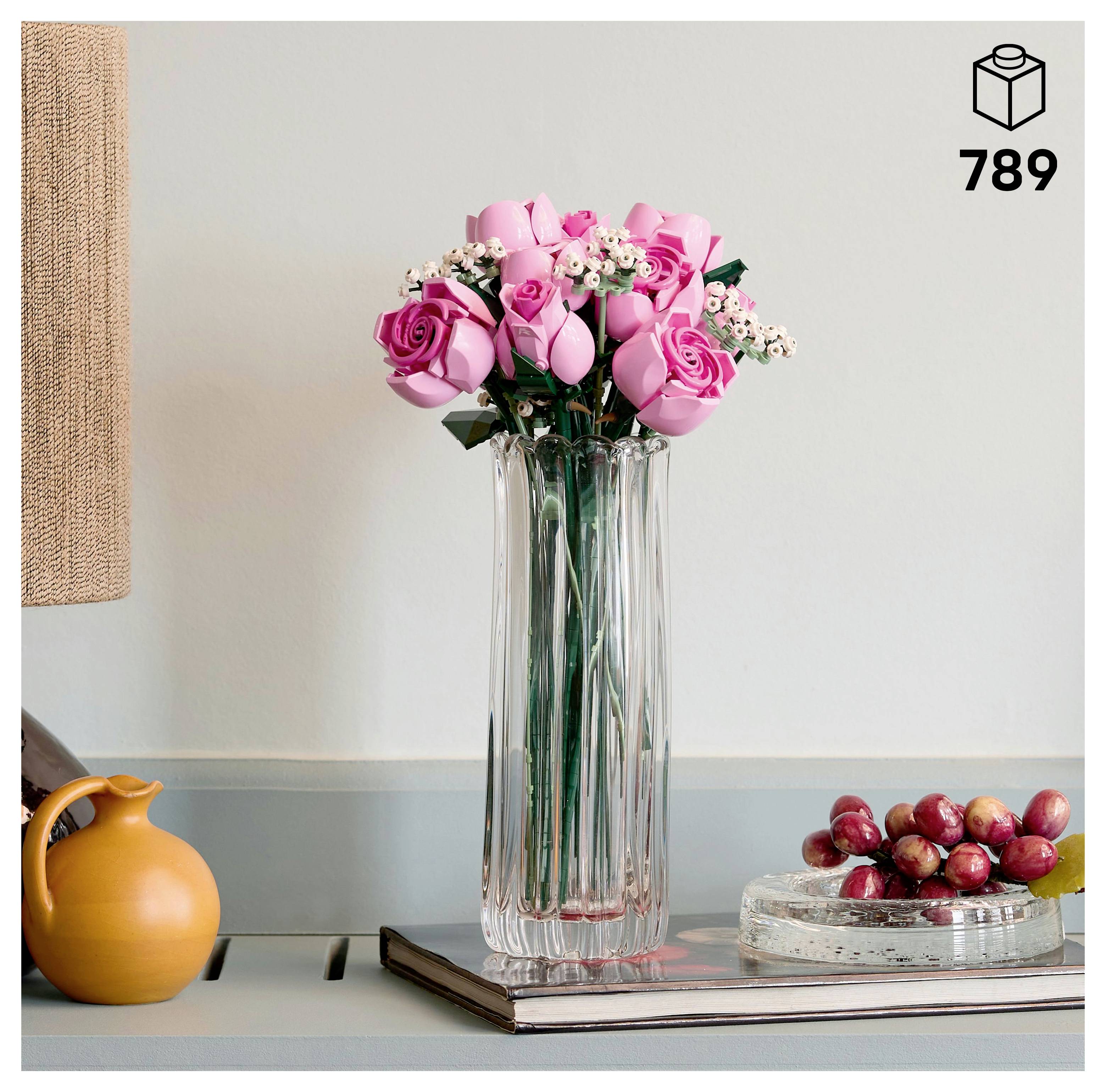 Un vase en verre transparent contient des roses roses et des fleurs blanches sur un livre. Un pichet jaune et un petit plat avec des raisins sont disposés à proximité sur une étagère.