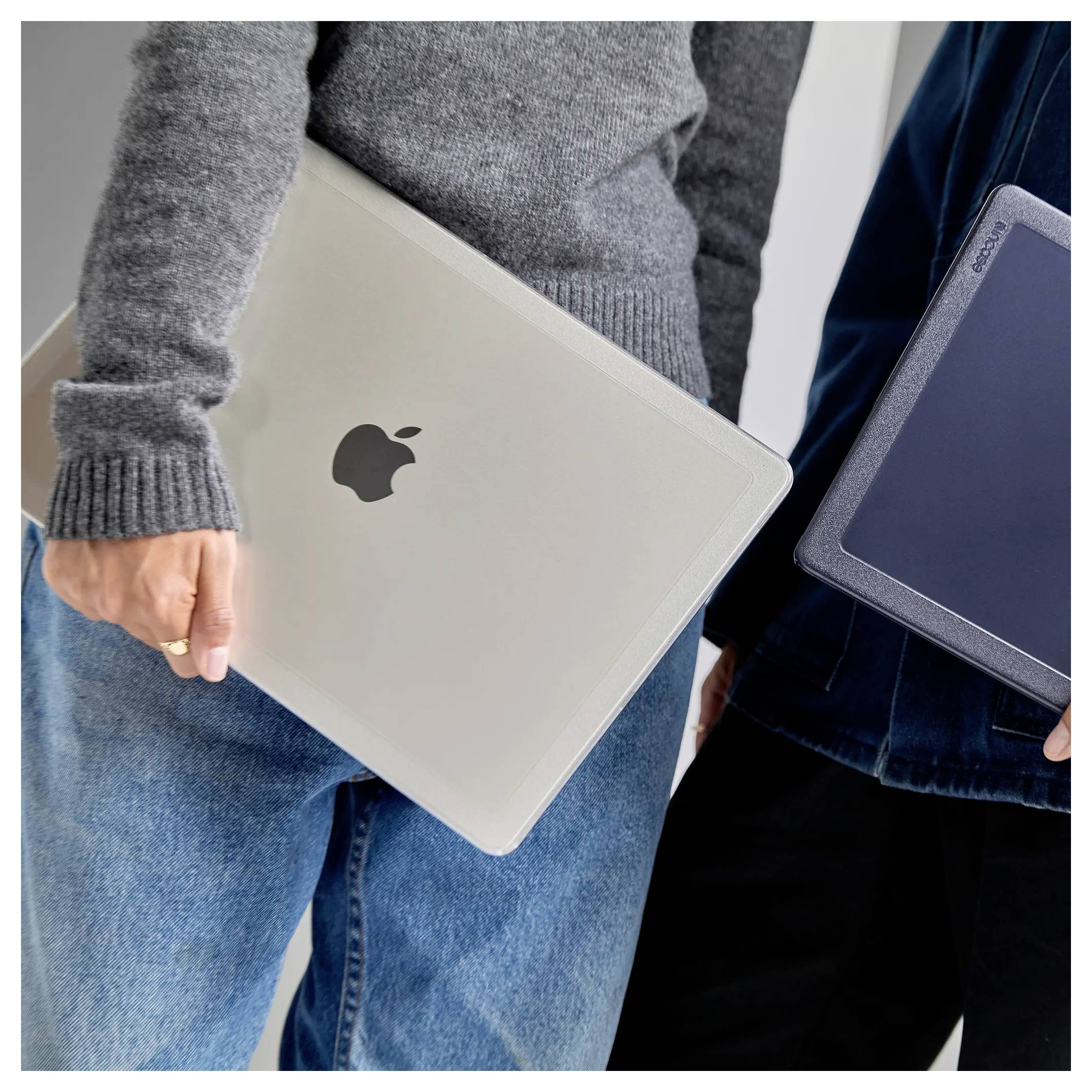 Une personne tient un ordinateur portable argenté fermé avec un logo Apple à côté d'une autre personne qui a une tablette. Les deux sont habillés de manière décontractée.