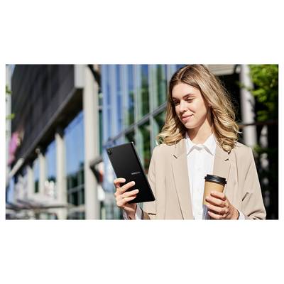Une femme se tient dehors devant un bâtiment moderne, tenant une tablette et une tasse de café, souriante et regardant l'écran.