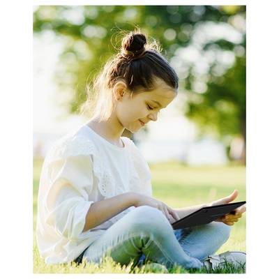 Une jeune fille est assise sur l'herbe dans un parc, concentrée sur une tablette. Elle sourit légèrement, la lumière du soleil filtrant à travers les arbres en arrière-plan.