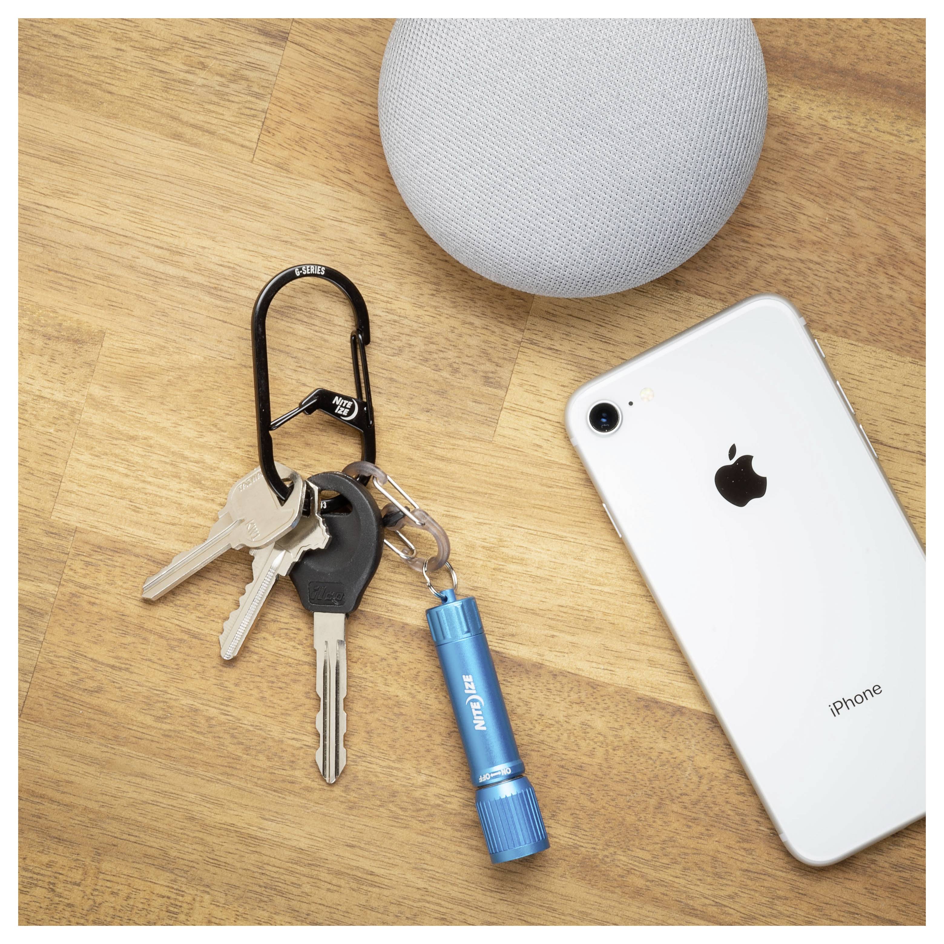 Des clés sur un mousqueton noir avec une lampe de poche bleue sont posées à côté d'un smartphone et d'un haut-parleur intelligent gris sur une surface en bois.
